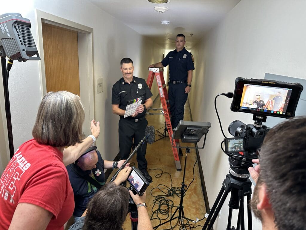 A film crew capturing audio and video footage of a man holding a clapper board and a man on a ladder both in public safety official uniforms.