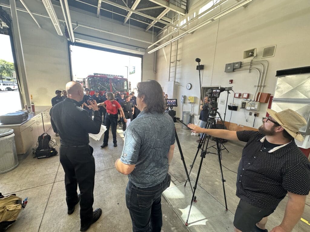 A film crew capturing audio and video footage of a fire department team in front of a fire engine parked outside its station.