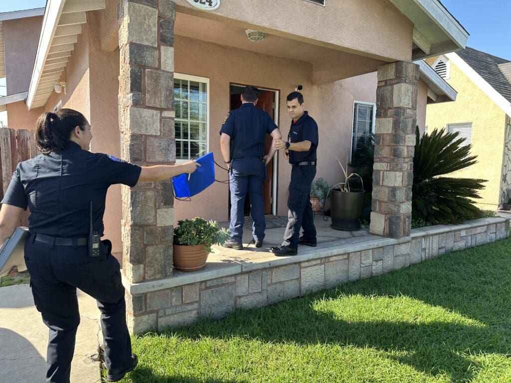A female public safety officer is handing her male colleague a piece of equipment while a third male colleague speaks to someone at the entryway of the home.