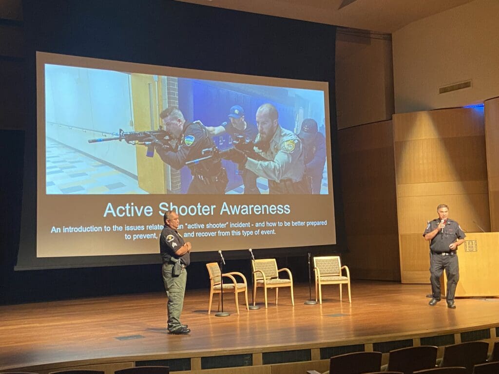 2 male members of MySafe:LA giving a presentation on active shooter awareness in an auditorium. A slide projected on the screen behind them shows an image of 4 men with guns and supporting text that reads "Active Shooter Awareness, an introduction to the issues related to an active shooter incident and how to be better prepared to prevent...and recover from this type of event."