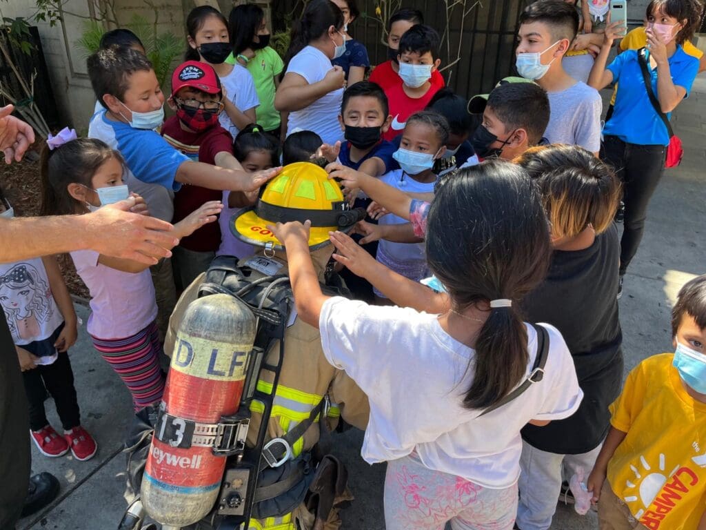 School children crowded around a crouched firefighter in full gear, allowing the children to touch his helmet. 