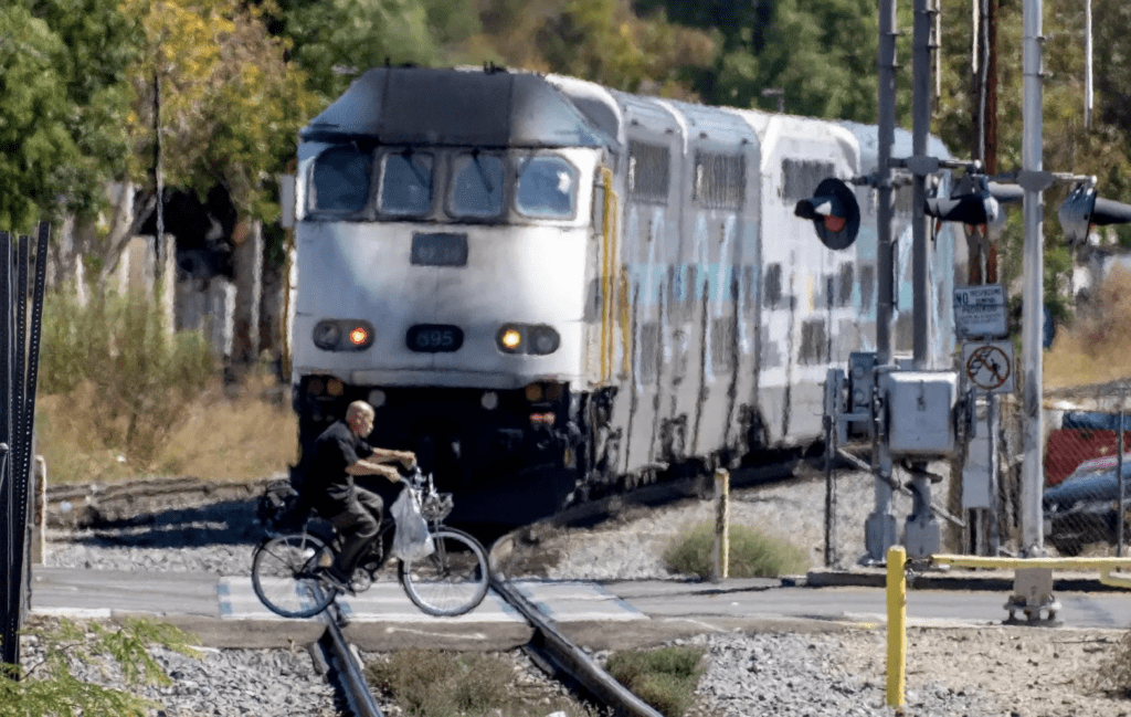 A silver train traveling along tracks while a man on a bicycle rides across the tracks in front of the incoming train.